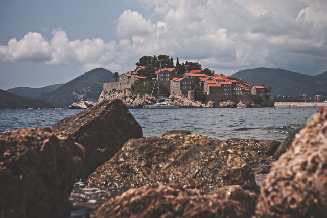 This beautiful scenery of a historical island village features charming stone cottages with characteristic red roofs. Surrounded by serene waters and rough rocky shores, with scenic clouds and hills in the background, the image radiates tranquility and beauty, perfect for travel guides, tourism promotional material, or landscape photography portfolios.