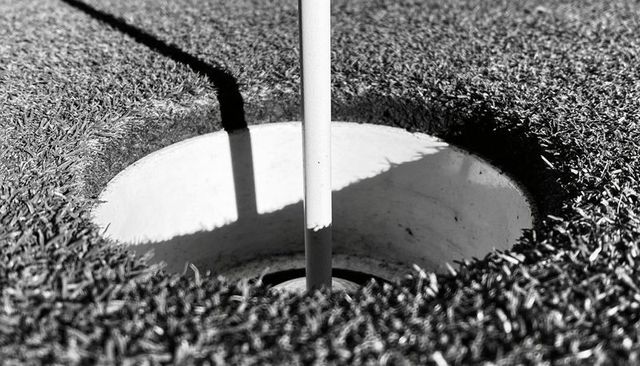 Close-up golf cup and flagstick casting diagonal shadow on putting green, monochrome