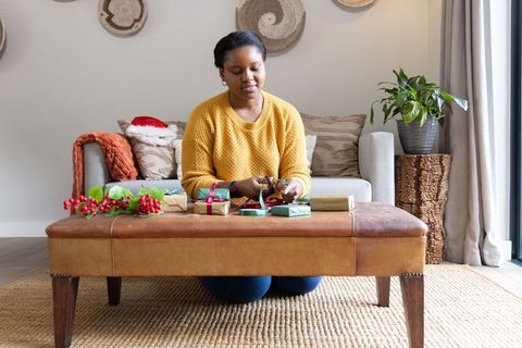 African american woman wrapping christmas gifts on ottoman in modern living room