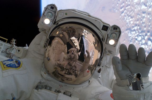 Image shows an astronaut in a space suit performing an extravehicular activity (EVA). The astronaut is waving, and another astronaut is visible in the reflection on the helmet, along with Earth's desert landscape approximately 225 miles below. This image can be used in publications or documentaries about space exploration, NASA missions, and science-related educational content.
