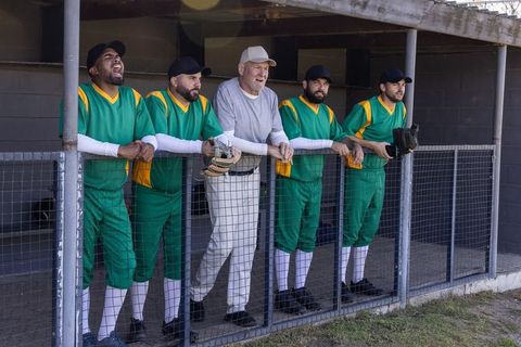 Veteran coach and diverse baseball teammates leaning in dugout cheering during game