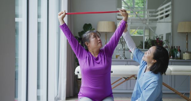 Senior woman practicing arm stretches with assistance from a physiotherapist using a resistance band in a living room. Great for use in articles or promotional materials about physical therapy, rehabilitation, elderly wellness, and home healthcare services.