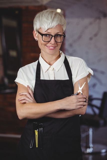 Female hairdresser with short hair and glasses standing confidently with arms crossed in a salon. She is wearing an apron and holding scissors, indicating her profession. This image can be used for promoting beauty salons, hairdressing services, or professional training courses in the beauty industry.