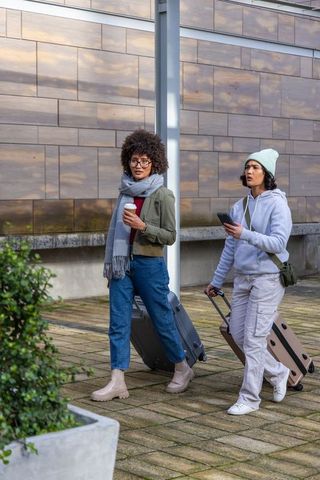 Two friends walking with rolling luggage and coffee in modern urban plaza, casual travel