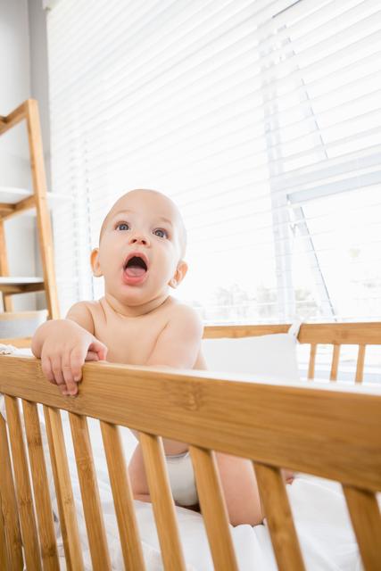 Baby boy playing in a wooden cradle at home with natural light streaming through the window. Ideal for use in parenting blogs, childcare articles, nursery decor advertisements, and baby product promotions.