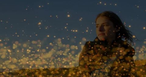 Contemplative woman standing at dusk with windblown hair and golden bokeh lights