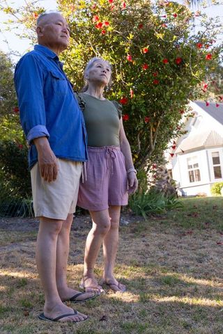Asian senior couple standing in sunlit suburban front yard near red flowering shrub