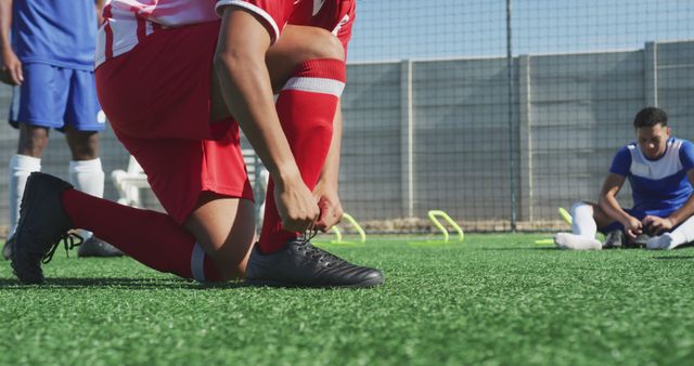 Focused soccer player kneeling and tying shoelaces on sports field while teammate stretches in background. Great for depicting sports preparation, teamwork, focus in sport activities and training routines.
