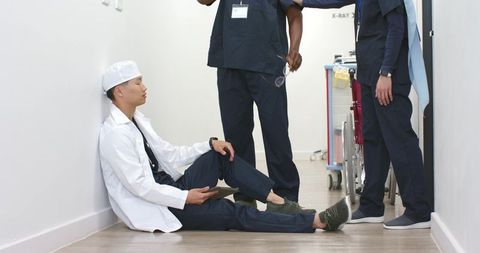 Asian physician sitting in corridor holding tablet with diverse medical team standing