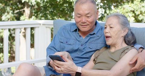 Senior asian couple sharing smartphone on sunny deck embracing technology and relaxation