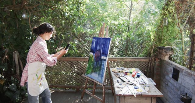 Female artist taking a photo of her painting in a serene garden studio. Perfect for websites and marketing materials related to art, creativity, hobbies, and outdoor activities. Also suitable for articles and content focusing on artistic inspiration and creative lifestyles.