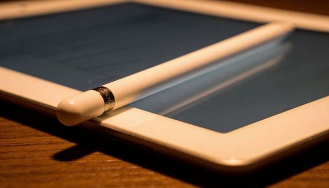 White stylus lying across tablet screen on wooden desk closeup with metallic band