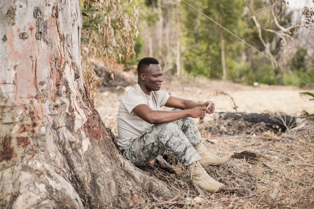Military soldier in camouflage uniform sitting under tree using mobile phone during training break. Ideal for content related to military life, communication technology, outdoor activities, and relaxation during training.