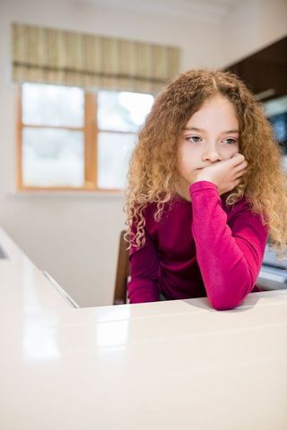 This image depicts a young child with curly hair sitting pensively at a shiny, white kitchen countertop. The scene is warmly lit with natural light streaming through a window with a cream and green valance, emphasizing the comfortable and modern home environment. This image is ideal for uses related to family lifestyle, home decor inspirations, and contemplative mood in editorial content.