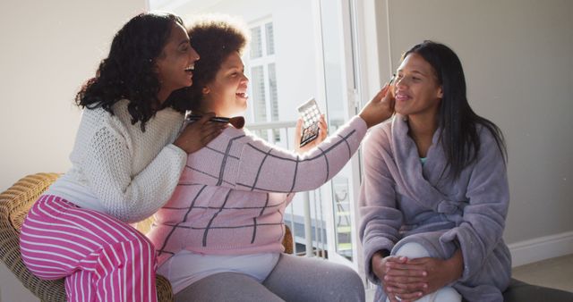 Three multiethnic women enjoying a fun makeup session at home. Women are smiling, bonding, and laughing while applying makeup. Perfect for use in promotions related to beauty, self-care routines, friendships, and diversity in lifestyle campaigns.