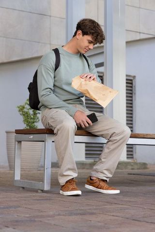 Male student sitting on campus bench holding lunch bag, smartphone wearing green sweatshirt