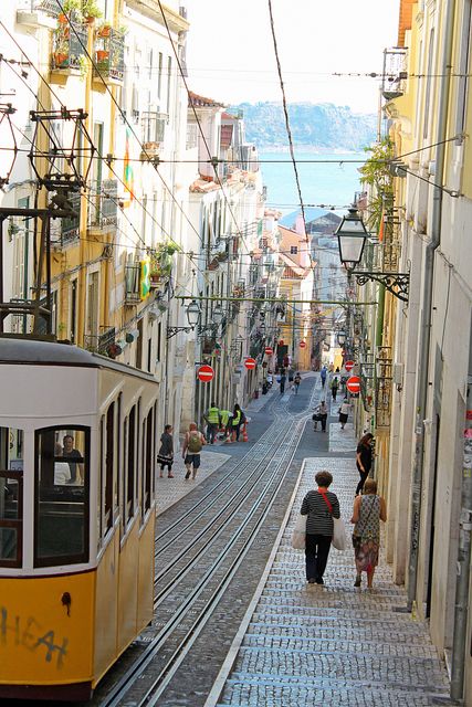 Lisbon tram moving up a cobblestone street surrounded by historic buildings with pedestrians walking. Ideal for travel blogs, cityscape presentations, and brochures marketing European destinations.