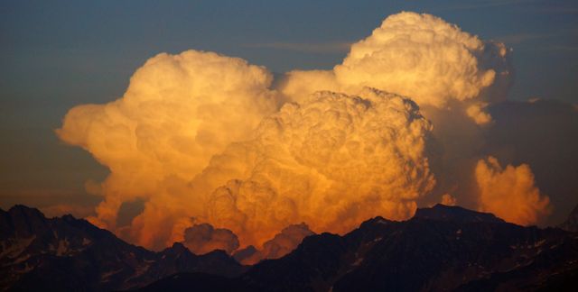 Beautiful cumulus clouds illuminated by the setting sun over silhouetted mountain range. Ideal for illustrating weather conditions, dramatic landscapes, and serene natural beauty in presentations or travel-related promotions.