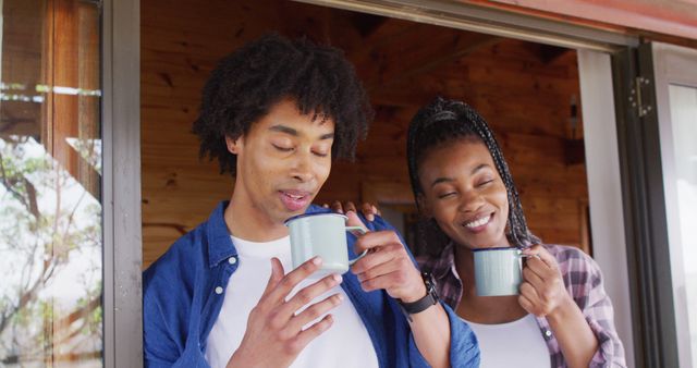 Happy african american couple looking through window and drinking coffee in log cabin, slow motion. Lifestyle, domestic life, countryside and nature concept.