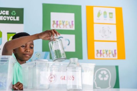 African american boy sorting glass jars into recycling bin in colorful classroom