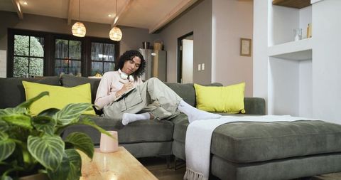 Non-binary adult relaxing on gray sectional sofa writing in journal wearing headphones