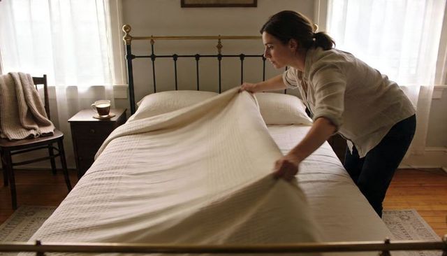 Woman making bed in sunlit bedroom with iron headboard, neutral linens, morning routine