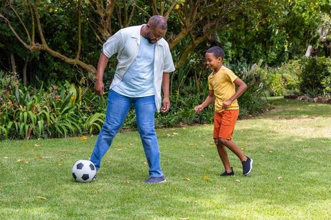 African american father playing soccer with son on backyard lawn in garden