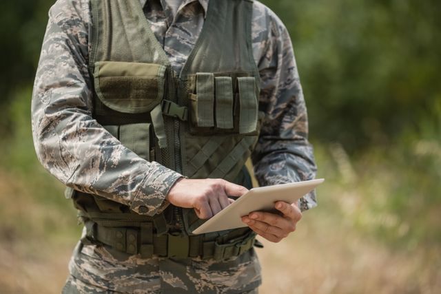 Mid-section view of military soldier interacting with digital tablet while in field. Useful for articles on modern military technology, tactical communications, defense strategies, and training methods.