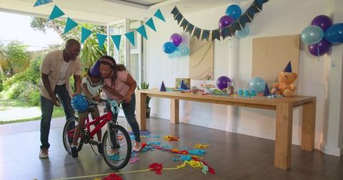 Diverse family helping child ride bicycle at home birthday party