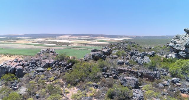 Photo showcases expansive rocky terrain with distant agricultural fields stretching to horizon. Clear blue sky enhances natural beauty. Perfect for themes on agriculture, rural living, geology, nature, outdoor adventure, environmental presentations, tourism advertisements, promoting countryside travel destinations, and illustrating traditional farming methods.