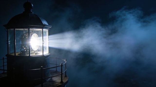 Lighthouse beacon piercing fog at night with fresnel lens beam on iron gallery