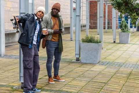 Two friends leaning in urban courtyard holding skateboard and coffee wearing beanies