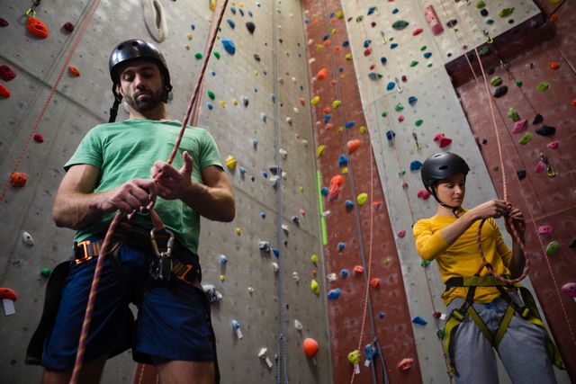 Low angle view of two athletes preparing for indoor rock climbing by tying ropes. Both are wearing safety gear including helmets and harnesses. The climbing wall in the background is equipped with colorful holds. This image is ideal for use in articles or advertisements related to fitness, sports, teamwork, and safety in climbing activities.