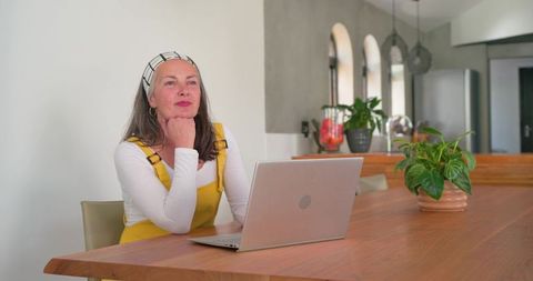 Woman wearing yellow pinafore gazing at laptop on wooden table for remote work inspiration