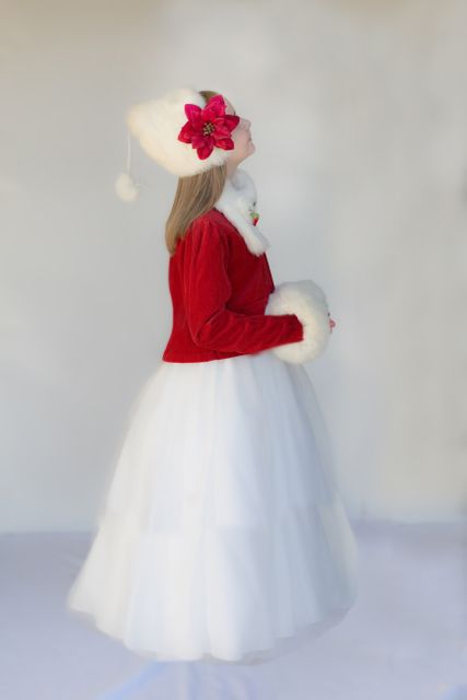 Young girl dressed in a festive red jacket with white fur lining and a matching white hat with a red flower and pompom. She is looking up, giving a side profile view as she wears a long white skirt. This image can be used for holiday greetings, festive fashion promotion, and winter clothing advertisements.