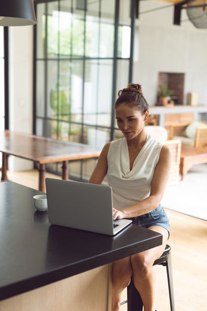This image depicts a woman sitting on a stool using a laptop on a kitchen worktop, representing a modern and relaxed home environment. Ideal for use in articles about remote work, home office setups, modern lifestyle, and technology use in daily life. It captures a casual and productive atmosphere, perfect for blogs, websites, and marketing materials targeting home-based professionals or lifestyle themes.