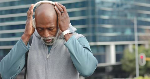 African american senior man adjusting white headphones on urban sidewalk by glass building
