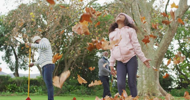 A family is spending time together in a park during autumn. A young girl is joyfully throwing leaves in the air, while two adults are engaged with raking. Lovely scene highlights bond, outdoor activities, and fall beauty. Great for articles about family bonding, seasonal outdoor fun, parks, and recreation.