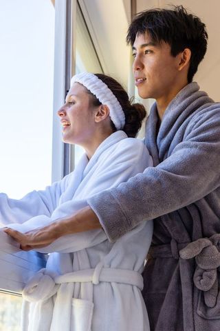 Diverse couple standing at window in plush robes enjoying sunny morning and cozy embrace