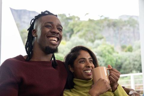 Happy couple relaxing on sofa smiling and holding mug by window with lush balcony view