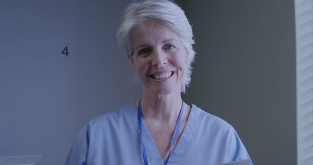 Senior female medical professional smiling in hospital corridor. Ideal for use in healthcare advertisements, medical website visuals, nursing recruitment materials, and promotional content showcasing hospital environments and medical staff. Can be used in content related to compassionate healthcare, patient care, or the healthcare industry workforce.