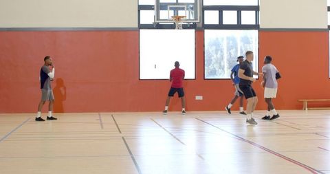 Five men playing pickup basketball on indoor gym court with hoop and daylight