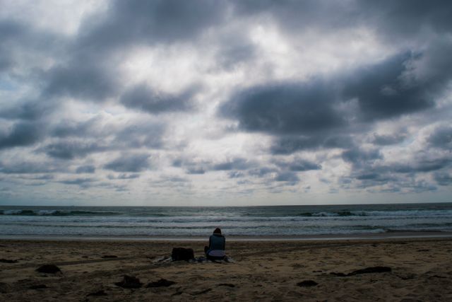 A single person sitting on a beach under a cloudy sky, looking at the ocean. Ideal for themes of solitude, contemplation, and moodiness. Can be used for stress relief promotions, meditation content, or artistic projects exploring loneliness and introspection.