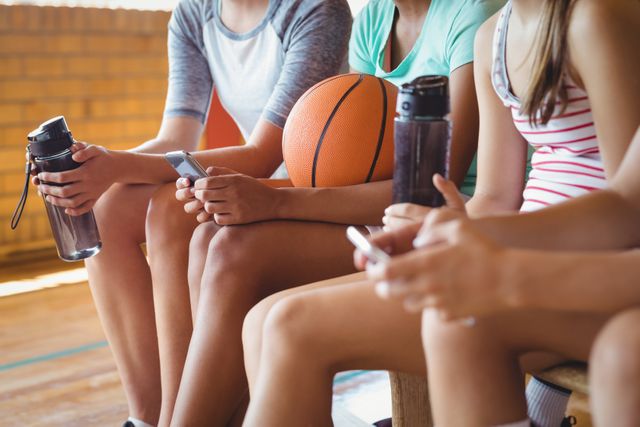 High school students sitting on a bench in a gymnasium, using mobile phones during a break from playing basketball. They are holding water bottles and a basketball is visible. This image can be used for themes related to youth, technology, sports, and social interaction.