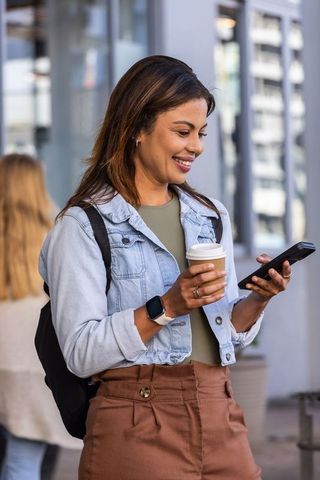 Young woman checking smartphone while holding coffee on urban sidewalk