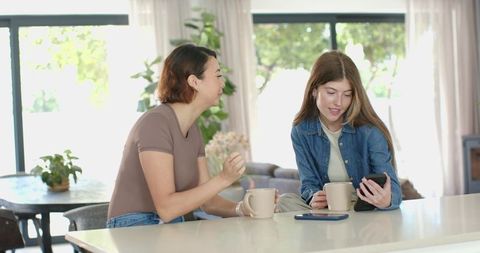 Female friends chatting over coffee at modern kitchen island, checking phone and laughing
