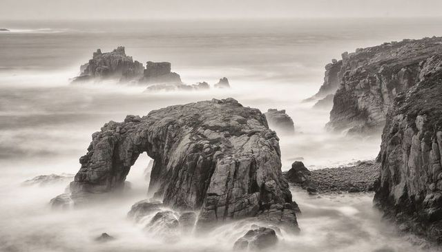 Weathered sea arch channeling mist and surf along rugged cliffside, monochrome coastal seascape