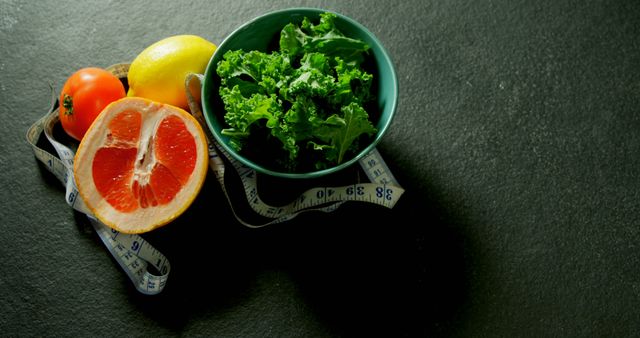 Depicts various healthy fruits and vegetables including a grapefruit, lemon, tomato, and a bowl of kale, with a measuring tape around them on a black background. Ideal for use in health, fitness, diet, and nutrition-related content, emphasizing the importance of a balanced diet and healthy lifestyle.