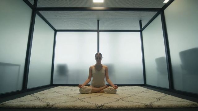 Office meditation woman meditating in minimalist glass-partition workspace on cream rug