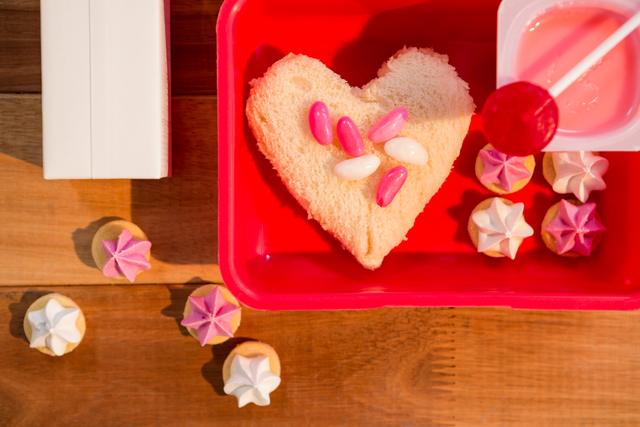 Heart-shaped sandwich with colorful candies and sweets in a red lunch box on a wooden table. Ideal for use in articles or advertisements about creative lunch ideas for kids, school lunches, or fun food presentations. Perfect for blogs, social media posts, or educational materials focusing on children's nutrition and meal planning.